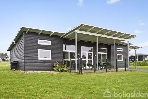 Et mørkt, moderne træhus med store vinduer og en overdækket terrasse. Huset ligger i et grønt, åbent område under en skyfri himmel.