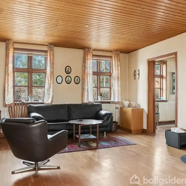 Stue med sort lædersofa og lænestol omkring et rundt bord på et tæppe. Brændeovn til højre. Lyse vægge og træloft med vinduer og gardiner i baggrunden.