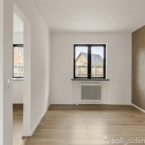 Et rum med en hvid væg og et stort vindue. Vinduet har udsigt til et hus. Trægulve og en væg med lodrette træpaneler skaber en minimalistisk atmosfære.