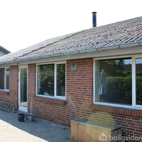 Rødt murstenshus har store vinduer og en skorsten, beliggende i en solrig gårdhave med flisebelagt terrasse. Træmøbler og grønne planter skaber en hyggelig atmosfære.