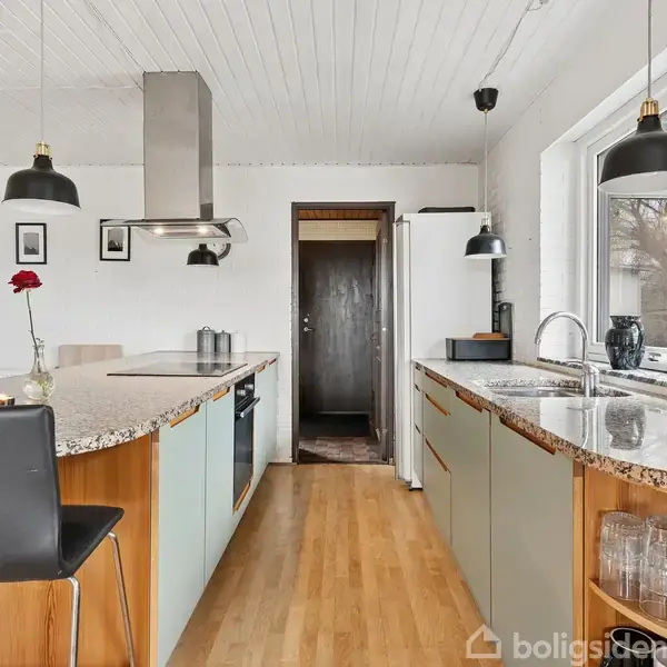 Køkken med grå skabe og en køkkenø med marmorbordplade. En sort stol står ved øen. To sorte pendellamper hænger fra det hvide loft. Vindue til højre.