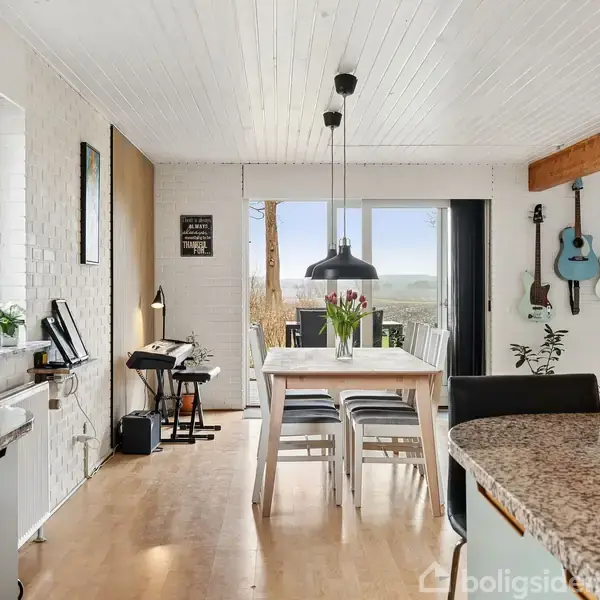 Spisebord med blomster står i en lys spisestue. Der hænger guitarer på væggen, og store vinduer giver udsigt til naturen udenfor. En lampe hænger over bordet.