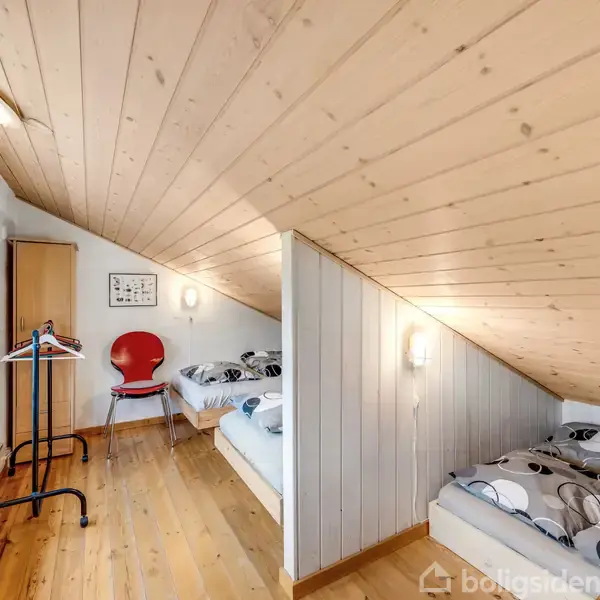 Et skrånende loftsværelse med trægulv har to senge i hver sin ende. En rød stol står langs væggen ved siden af et bord med en lampe. Værelset har lyse vægge og træloft.