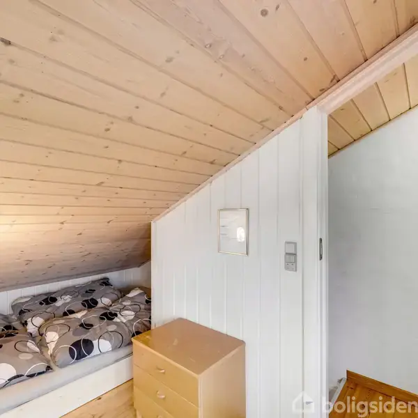 Seng placeret under et skråt loft med moderne, mønstret sengetøj; ved siden af står en lille kommode. Væggene er træpanelerede, og en gang fører videre mod et andet rum.