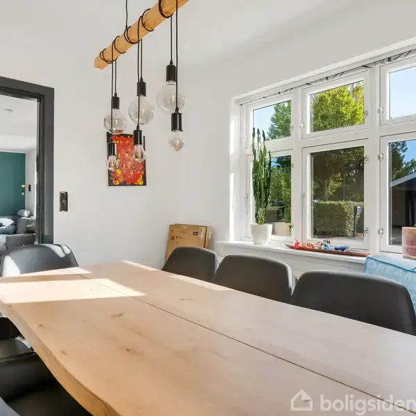 Spisebord af træ med sorte stole omkring, under hængende lamper. Til venstre åbner en dør ind til stue med sofa. Vindue til højre viser grøn udendørs have.