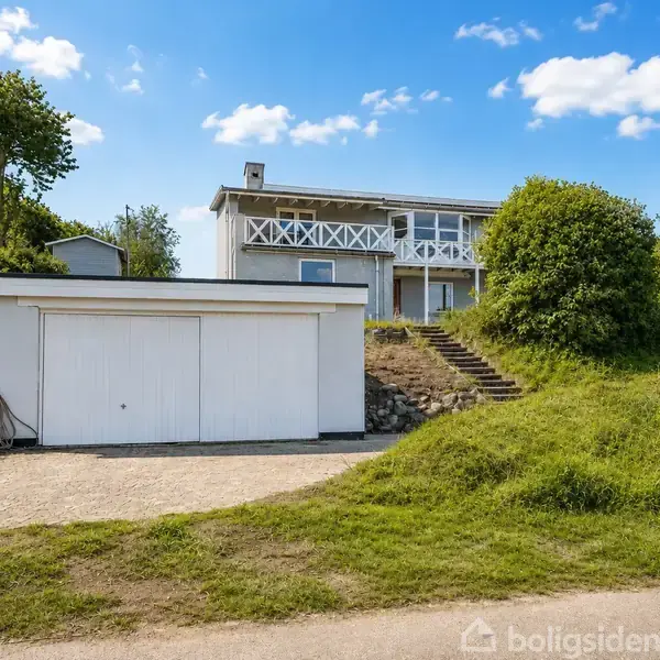 Et hvidt hus med en balkon står på en forhøjning. Foran er en garage og en grussti. Et skilt med teksten "Til salg" står til højre, omgivet af grønt landskab.
