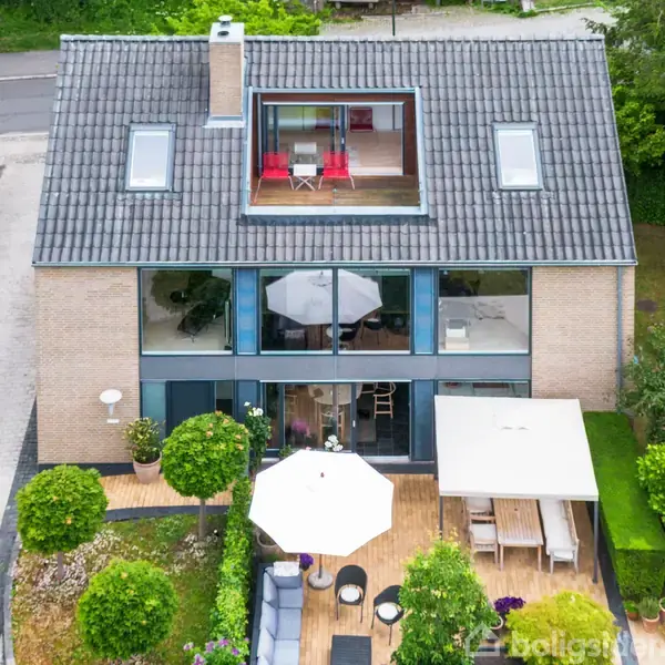 Et to-etagers hus med fladt tag, store vinduer og en møbleret terrasse med to parasoller i en grøn have, ligger ved siden af en asfaltvej omkranset af hække.