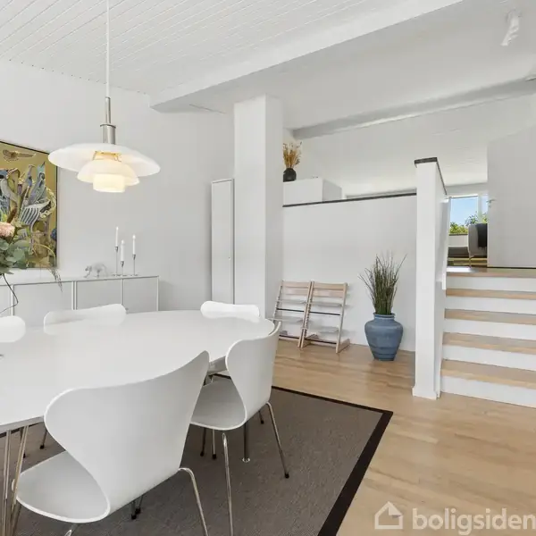 Et moderne spisestuemiljø med et ovalt hvidt bord omgivet af matchende stole. Bordet er dekoreret med en blomsterbuket. Rummet har lyse trægulve og en malet vægkunst.