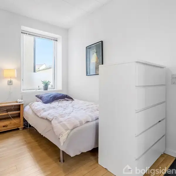 En enkeltseng med hvidt sengetøj står op ad en væg. Ved siden af er en trækommode og et vindue. En hvid skuffekommode står ved en dør. Væggen er dekoreret.