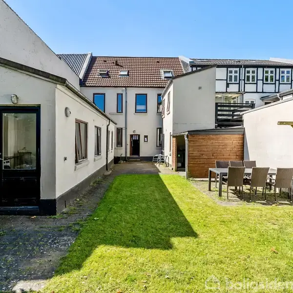 Baggård med grøn græsplæne og havemøbler af træ ved et bord. Bygninger på begge sider med vinduer og døre, under en klar blå himmel.