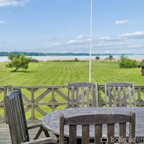 Et rundt havebord med stole står på en træterrasse. I baggrunden ses en stor, grøn græsplæne, træer og en sø under en klar blå himmel.