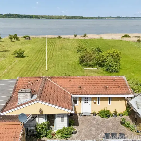 Et gult hus med rødt tegltag står ved en stor, velplejet græsplæne. I baggrunden ses en strandbred ved en sø eller fjord omgivet af frodig natur og blå himmel.