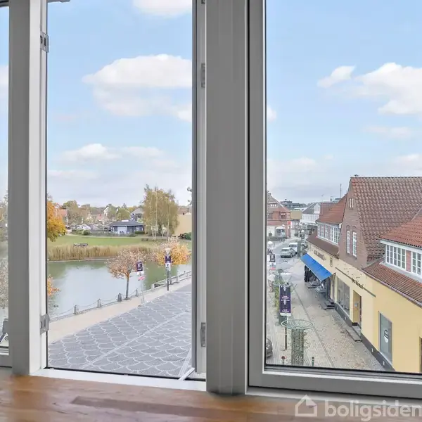 Et åbent vindue viser udsigt til en stille gade med gule bygninger og en flod i nærheden. Der er træer og klar blå himmel med enkelte skyer i baggrunden.