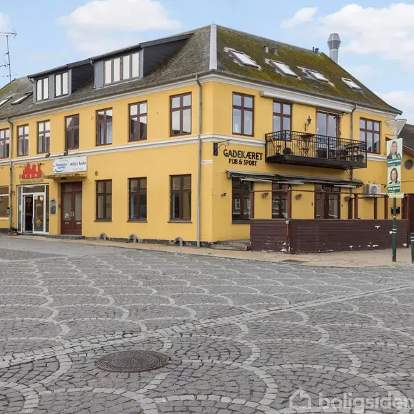 Et funktionelt køkken med sort-hvidt skakternet gulv. Induktionskomfur og vask under skråvæg med ovenlysvinduer. Lyse skabslåger og hylder nær en radiator.