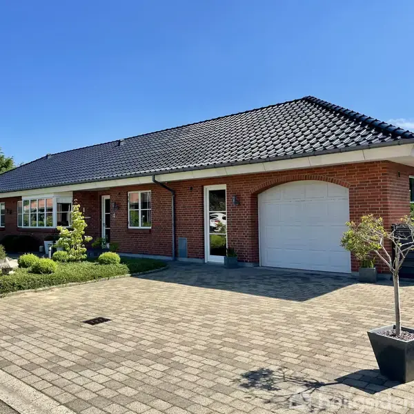 En rød murstensvilla med sort tegltag står omkranset af en velplejet have. En flisebelagt indkørsel fører til garageporten. Grønt løv og himmel danner baggrund.