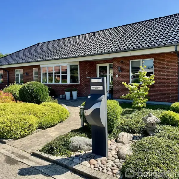Rød murstensbungalow har flisebelagt indkørsel og velplejet have med buske og sten. En postkasse viser “Borger & Tofte 4 Lykkevej” foran huset i et villakvarter.