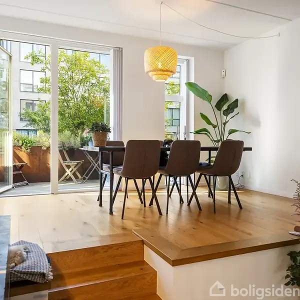 Et spisebord med fire stole står i et lyst rum med trægulv. Store glasdøre fører ud til en altan med planter, mens en gul lampe hænger over bordet.