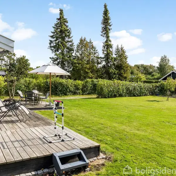 En træterrasse med stole og parasol står ved et hus. Foran er en velplejet græsplæne omkranset af buske og træer, der giver en fredelig atmosfære.