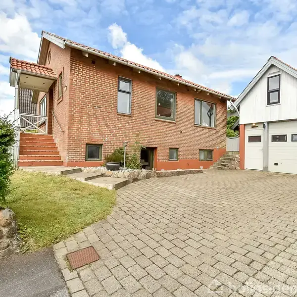 Rødt murstenshus med tre trapper foran ses ved siden af en grå garage med en sort affaldsbeholder. Brostensbelagt indkørsel fører til begge bygninger i et beboelsesområde. Nummer 10 vises.