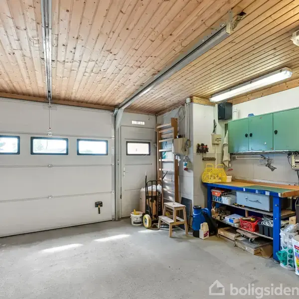 Et ryddeligt værksted med arbejdsbord står langs en væg med ophængt værktøj og grønne skabe. Garageporten har små ruder, og rummet er oplyst af loftslamper.