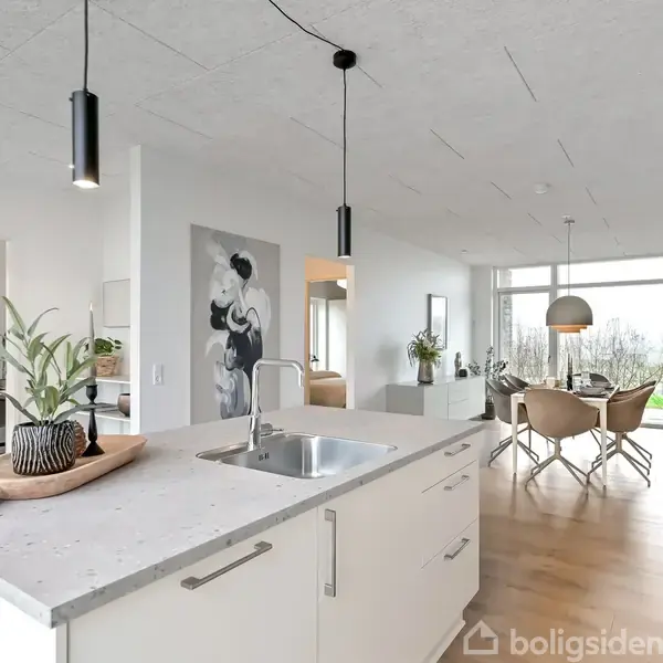 Køkkenø med moderne design står centralt med en grå bordplade og vask. Rummet er åbent, lyst, med trægulve og spisestue i baggrunden. Billede på væggen.