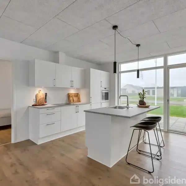 Et moderne køkken med en hvid køkkenø og to sorte barstole. Køkkenet har indbyggede hvide skabe og apparater. Store vinduer kigger ud mod en åben græsplæne.