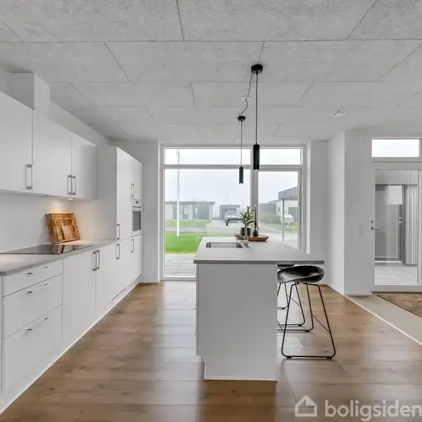 Et moderne køkken med hvide skabe og en central ø, to sorte barstole står ved siden af. Store vinduer overser en grøn græsplæne, der fylder baggrunden med naturligt lys.