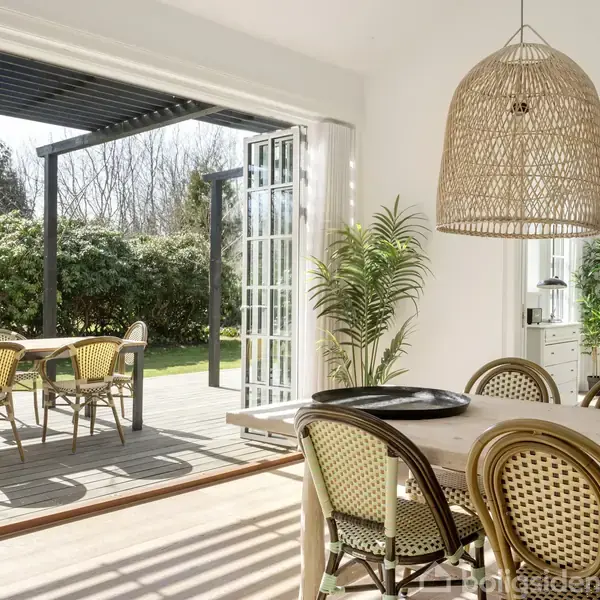 Fletkurvslampe hænger over spisebord i lyst rum, som åbner op til terrasse med havemøbler. Grønne planter omkranser området, derudover er der sollys, der strømmer ind.