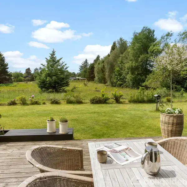En terrasse med flettede stole og et bord med en kaffekande og aviser. I baggrunden er en stor, grøn have med træer og buske under en blå himmel med skyer.