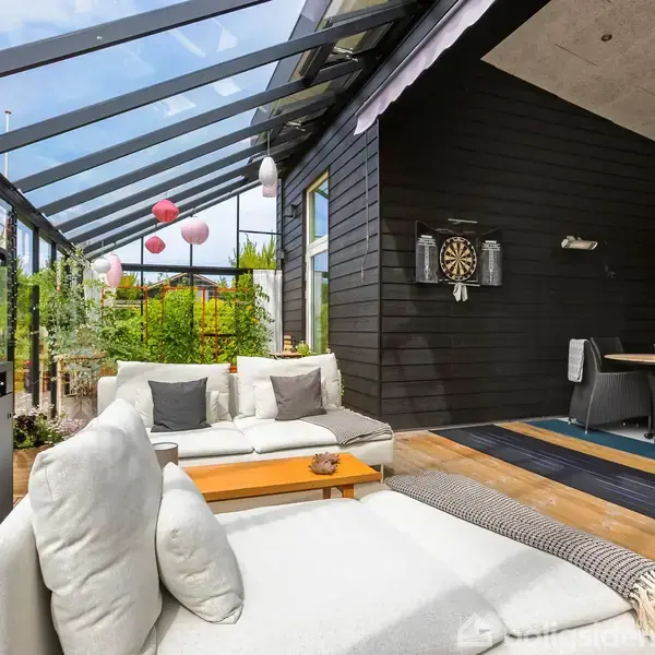 Sofaen står i en overdækket, udendørs stue med gennemsigtigt tag. En dartskive er monteret på en sort væg. Der er også en terrassevarmer og flere lanterner i baggrunden.