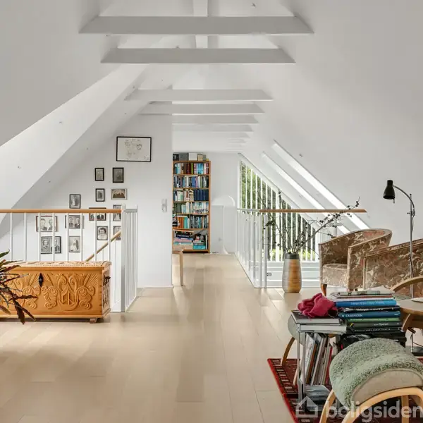 Stue med loftbjælker indeholder loungestole, et bord med bøger og en lampe. Store vinduer lukker lys ind, omgivet af bogreoler og stueplanter, giver et lyst og hyggeligt rum.
