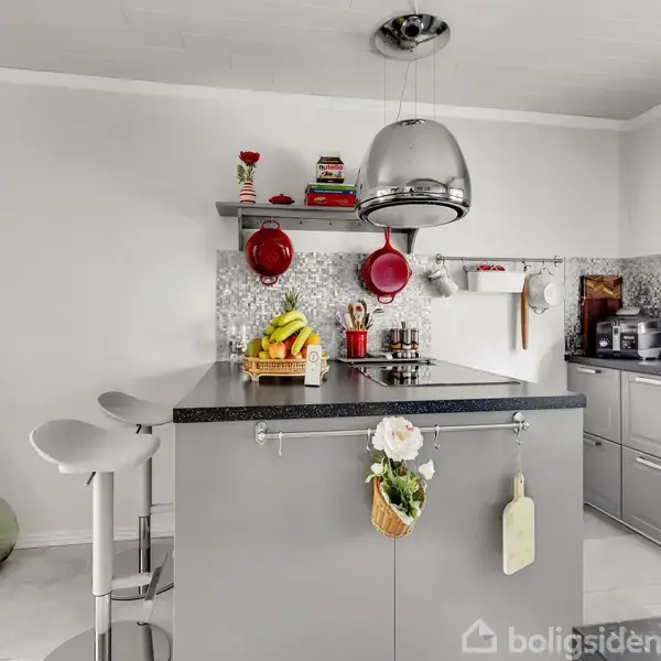 Et moderne køkken har en grå køkkenø med frugtskål og hængekrog. Barstole står ved siden af, og der hænger en stor lampe ovenfor. Væggene er hvide med hylder og dekorationer.