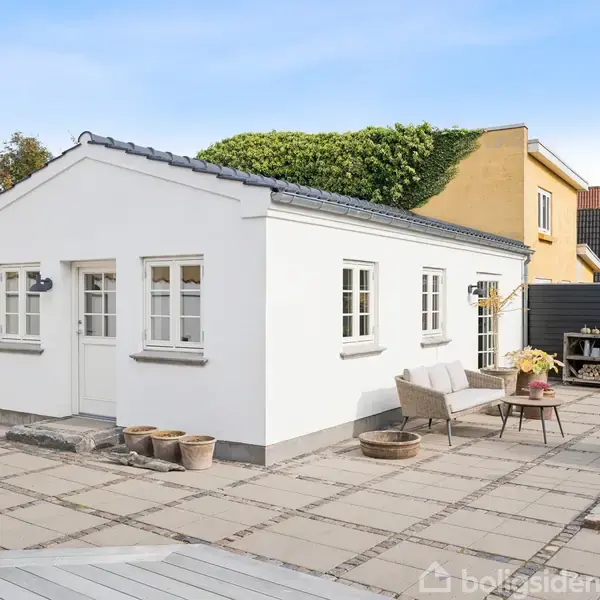 Hvid bygning med flere små vinduer står stille på flisebelagt terrasse. Omkring er der havemøbler og blomsterpotter. Baggrunden viser flere nabohuse og blå himmel.