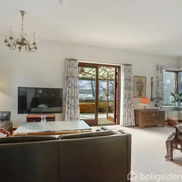 Sofaer og lænestol står i en stue med en fladskærms-tv på et møbel. Stuen har lyse farver, et vindue med udsigt og en dør til en terrasse, omgivet af diverse nips og malerier.