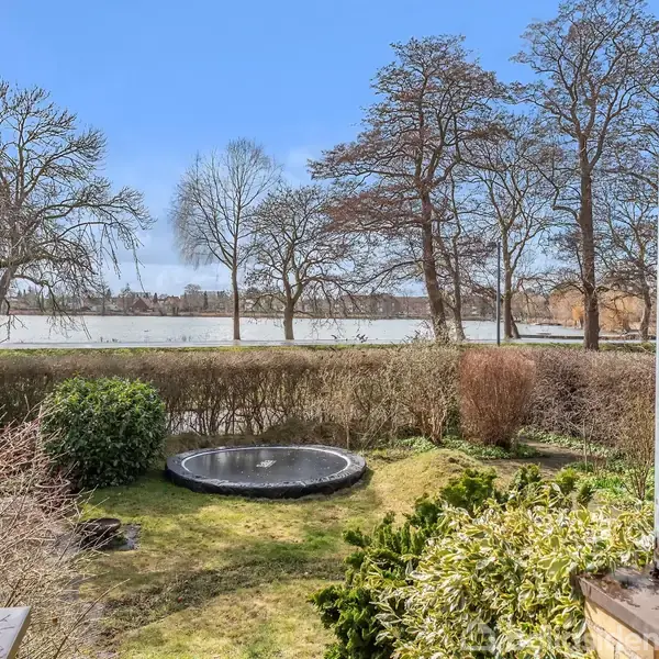 Trampolin i haven omgivet af græsplæne og buske, med kig ud over en sø og høje træer i baggrunden under en klar blå himmel.