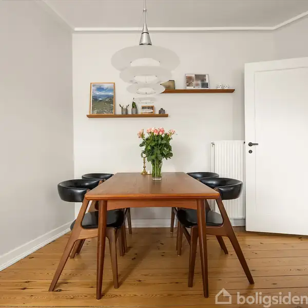 Et træbord med en blomsterbuket står omgivet af seks sorte stole. Rummet har trægulv, hvide vægge, et maleri, og en hylde med små dekorationer.