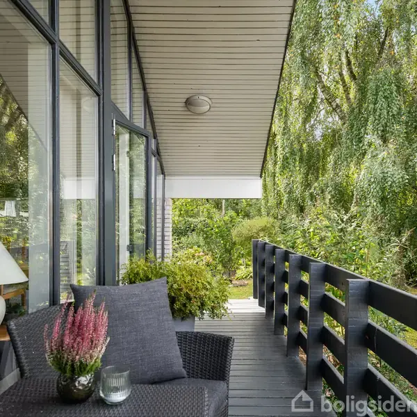 En terrasse med sorte møbler og en plantekrukke står ved siden af et hus med store glasvinduer. Omgivet af frodig vegetation, træer og en græsplæne.