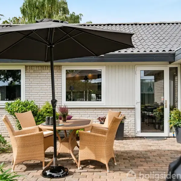 Terrassemøbler står under en stor parasol, placeret på en stenterrasse foran et hvidt murstenshus. Planter omkranser området, mens en glasdør fører ind i huset.
