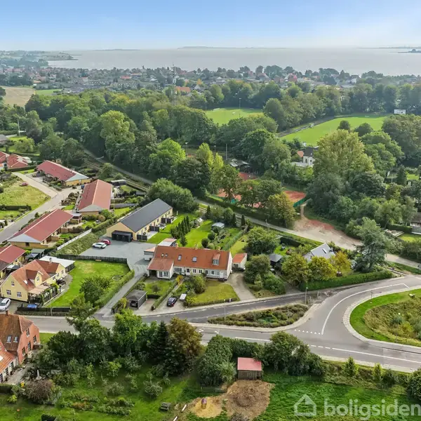 Landsby med huse omgivet af grønne områder og veje. I baggrunden ses skov og en sø eller fjord. Boligerne har haver og ligger tæt på hinanden.