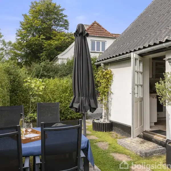 Terrassemøbler står omkring et bord dækket med en blå dug og en ubrugt parasol. Bordet står ved siden af et hvidt hus i en have omgivet af grønne hække.