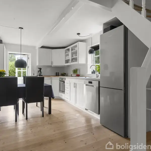 Et moderne køkken med hvide skabe og grå hårde hvidevarer; et spisebord med sorte stole står ved et vindue, ved siden af en trappe med hvide balustre.