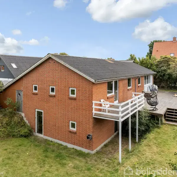 Rødt murstenshus med sort tag står stille i en grøn have. En trappe fører til en hævet træterrasse med stole. Omgivet af tætte buske og træer.