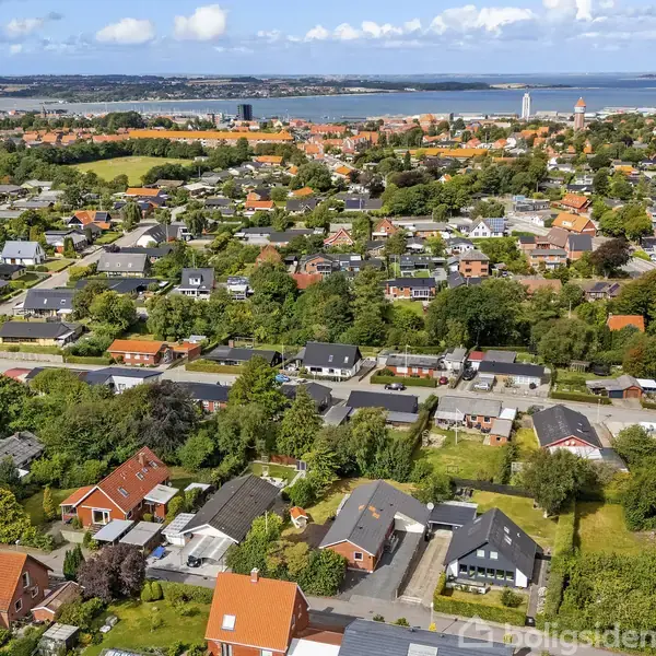 Boligområde ses fra luften med røde og sorte tage under spredte skyer. I baggrunden er der en by ved en kystlinie, omgivet af træer og marker.