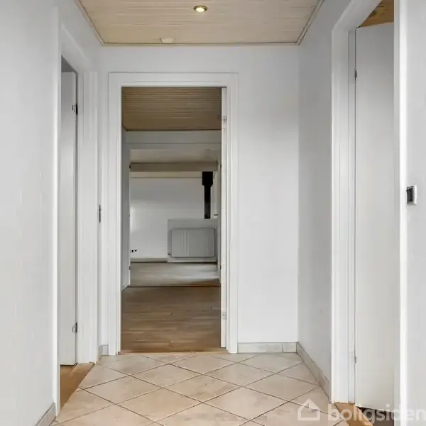 En lys gang med gulvfliser leder til en åben dør, der fører ind i et rum med trægulv og hvidmalede vægge. To døre findes på hver side.