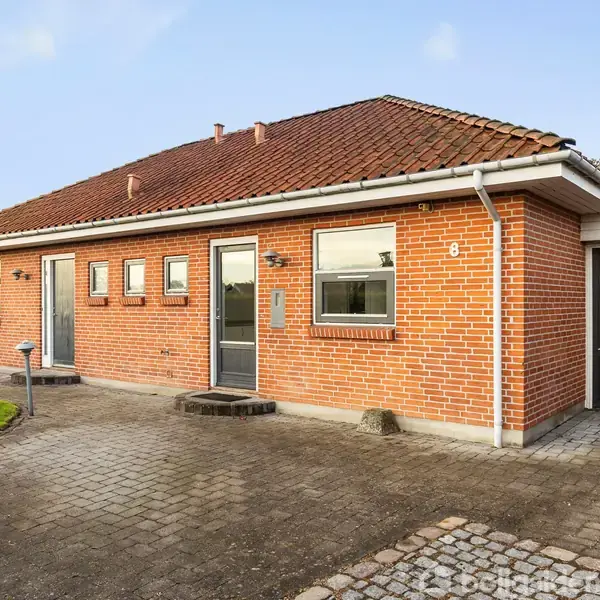 Rødt murstenshus står stille ved flisebelagt indkørsel i et villakvarter. Tre vinduer og to døre på facaden. I venstre side er en grøn græsplæne. Nummeret "8" er på facaden.