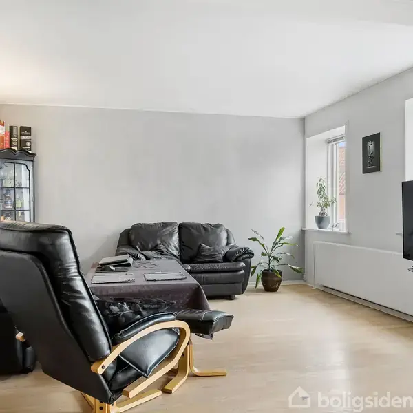 En stue indeholder sorte lædersofaer omkring et sofabord. En fjernsynsskærm står ved vinduet, og en plante og billeder dekorerer væggene. Rummet er lyst med parketgulv.