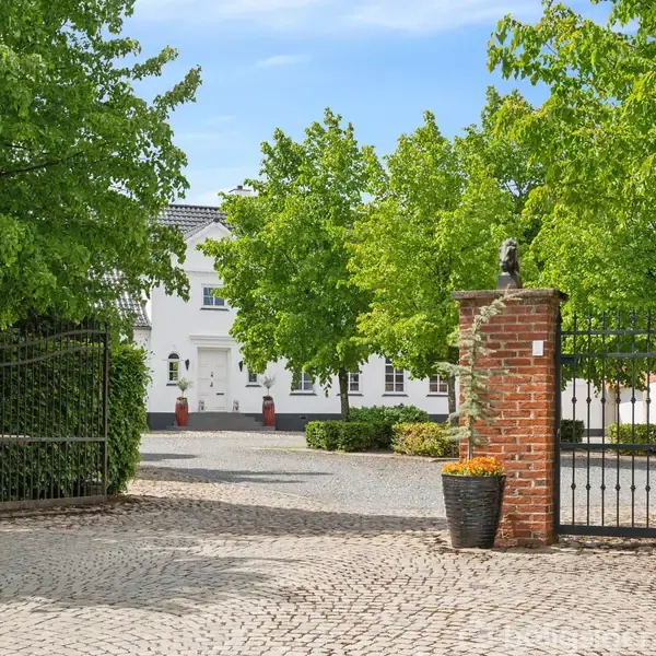 En åbent smedejernslåge pryder en brostensbelagt indkørsel omgivet af murstenssøjler med blomsterkrukker. Træer omkranser indgangen, og i baggrunden ses et hvidt hus. Skilt med tallet 24.