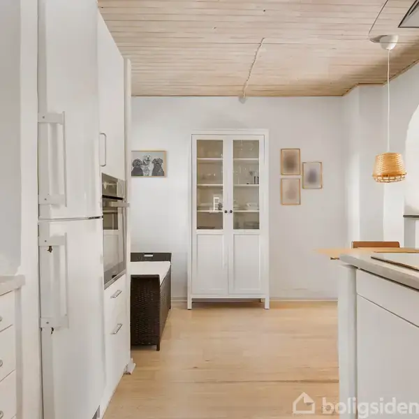 Køkken med hvide skabe og indbygget ovn, hvor en emhætte hænger over kogepladen. I baggrunden ses en hvid vitrine og vægdekorationer mod en lys trævæg og trægulv.