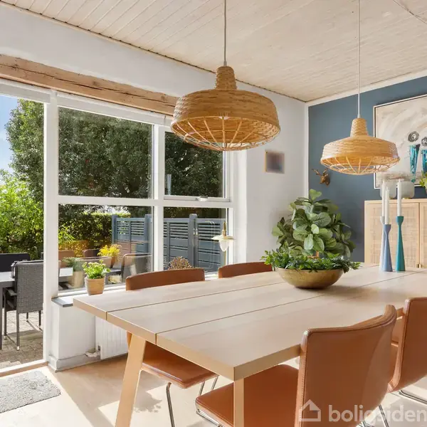 En spisestue med et træbord omgivet af brune stole, hvorpå der står en skål med en plante. To lamper hænger over bordet. Dørene åbner til en terrasse med havemøbler udenfor.