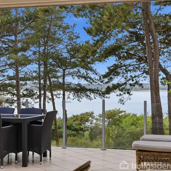Havemøbler står på en terrasse med udsigt til en skov og vand. Træer omgiver området, og blå himmel ses i baggrunden.